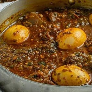 May include: A close-up shot of a stainless steel pot filled with a stew. The stew is a rich brown color with visible green herbs and several golden-yellow hard-boiled eggs. The dish appears to be freshly cooked and ready to serve.