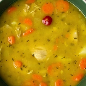 May include: A close-up shot of a bowl of soup with a vibrant yellow broth. The soup contains visible pieces of orange carrots, white vegetables, and a single red berry. The soup is garnished with dark green herbs and spices.