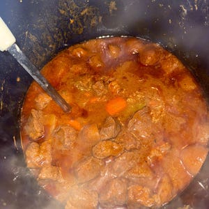 May include: A dark blue pot filled with beef stew. The stew contains large pieces of meat, carrots, and other vegetables in a rich, brown gravy. Steam rises from the hot stew, and a spoon rests in the pot.