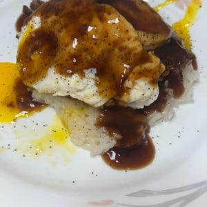 May include: A plate of food featuring a fried egg with a runny yolk, drizzled with a dark brown sauce. The egg sits atop a bed of white rice and a darker, meat-like substance. The plate has a floral design.