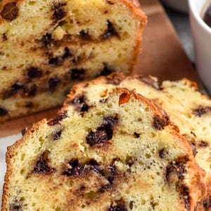 May include: A loaf of chocolate chip bread, partially sliced, with visible chocolate chips throughout. The bread has a golden-brown crust and a soft, yellow interior. Several slices are arranged in front of the loaf, showcasing the texture and chocolate distribution.