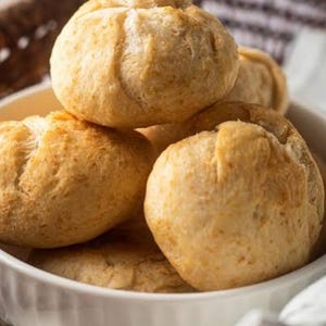 May include: A close-up shot of several golden-brown, freshly baked rolls in a white, scalloped-edge bowl. The rolls have a rounded shape with a textured surface, suggesting a soft interior. The image evokes a sense of warmth and homemade goodness.