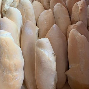 Puede incluir: Primer plano de varios panes recién horneados. Los panes, de color marrón claro, son de forma oblonga y tienen una textura ligeramente rugosa. Algunos panes revelan un interior pálido y cremoso. Un alimento básico.