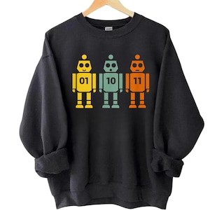 May include: Black sweatshirt featuring three colorful robot figures. Each robot is a different color: yellow, teal, and orange. The robots have the numbers 01, 10, and 11 printed on their chests. The sweatshirt has a crew neck and long sleeves.
