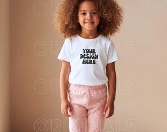 May include: A white t-shirt with the text "YOUR DESIGN HERE" in black, paired with pink pajama pants with white polka dots. The child is smiling, with curly brown hair.