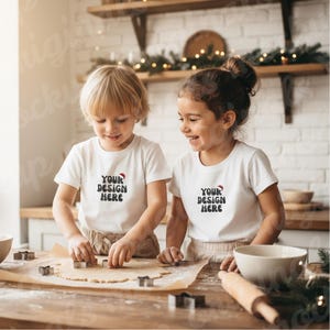 May include: Two children wearing white t-shirts with the text "YOUR DESIGN HERE" and a Santa hat graphic are smiling while cutting out cookies. The scene takes place in a kitchen with a wooden countertop and shelves.