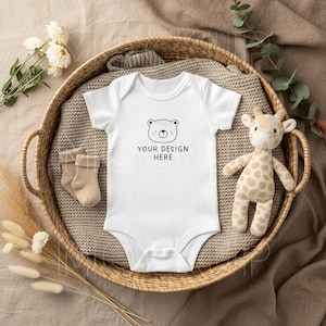 May include: A white baby bodysuit with a bear face graphic and the text "YOUR DESIGN HERE" is displayed in a wicker basket with a stuffed giraffe, beige socks, and decorative plants. The bodysuit is short-sleeved.