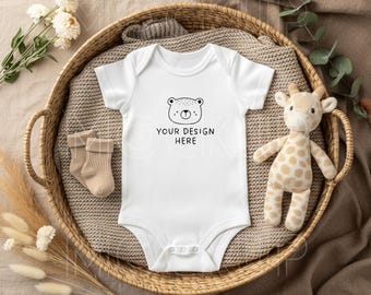 May include: A white baby onesie with a bear face graphic and the text "YOUR DESIGN HERE" is displayed in a wicker basket with a stuffed giraffe, beige socks, and decorative plants. The onesie is short-sleeved.