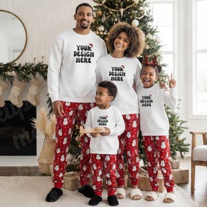 以下が含まれることがあります： お揃いのクリスマスパジャマを着た4人家族。白いスウェットシャツには黒字で「YOUR DESIGN HERE」と書かれています。赤いパジャマパンツには白い雪だるま模様。家族は、装飾されたクリスマスツリーの前に立っています。