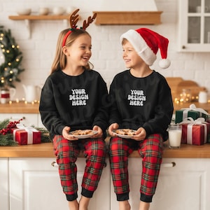 May include: Two children wearing matching black sweatshirts with the text "YOUR DESIGN HERE" and red plaid pajama pants. One child wears a Santa hat, the other a reindeer antler headband. They are holding plates of cookies, with gifts and a glass of milk nearby.