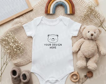 May include: A white baby onesie with a bear face design and the text "YOUR DESIGN HERE". The image includes a plush teddy bear, a wooden rainbow, baby shoes, and a wooden teether, all in neutral tones.