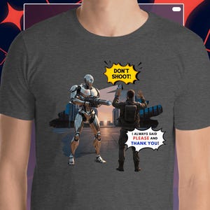 Op de afbeelding: Donkergrijs t-shirt met een afbeelding van een robot die een wapen op een persoon richt met de handen omhoog. Tekstballonnen zeggen "DON'T SHOOT!" en "I ALWAYS SAID PLEASE AND THANK YOU!"