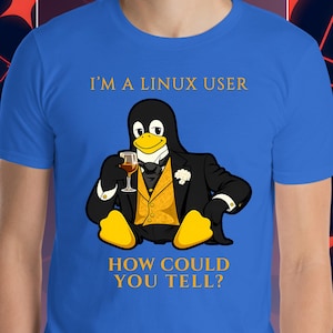 May include: Royal blue t-shirt featuring a tuxedo-clad penguin holding a glass of wine. The penguin has yellow feet and a yellow vest. Text on the shirt reads, "I'M A LINUX USER HOW COULD YOU TELL?"