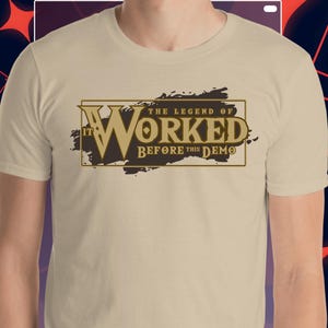 Pode incluir: Camiseta bege com o texto "IT WORKED The Legend of Before This Demo" em um design marrom e dourado. A camiseta de manga curta parece ser feita de um material macio.