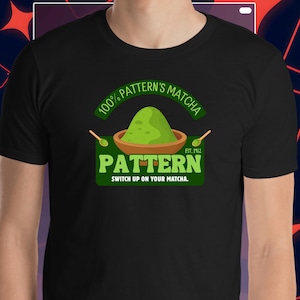 Puede incluir: Camiseta negra con un diseño de matcha verde. El diseño incluye el texto "100% Pattern's Matcha" y "Pattern", con el lema "Switch up on your matcha". También se muestra un cuenco de matcha y dos cucharas.
