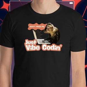 Könnte beinhalten: Schwarzes T-Shirt mit einer Grafik eines Affen, der einen Laptop benutzt. Der Text auf dem Shirt lautet "DON'T MIND ME" über dem Affen und "Just Vibe Codin'" darunter. Der Affe ist braun und beige.