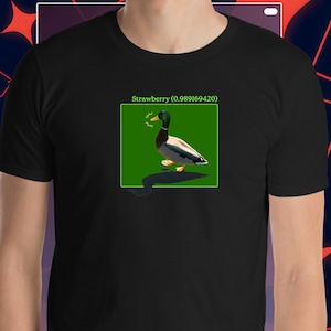Könnte beinhalten: Schwarzes T-Shirt mit einer Grafik einer Ente in einem grünen Quadrat und dem Text "Strawberry (0.989169420)". Die Ente hat einen grünen Kopf, einen braunen Körper und gelbe Füße. Das Shirt ist aus weichem Material.
