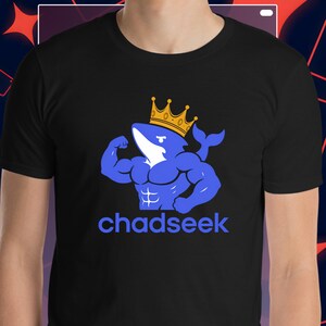May include: A black short-sleeved t-shirt featuring a graphic of a muscular blue shark wearing a yellow crown. The shark is flexing its arm, and the text "chadseek" is printed in blue letters directly underneath the graphic illustration.
