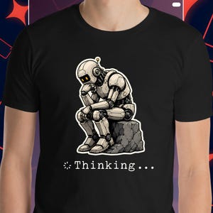 Può includere: T-shirt nera con una grafica di un robot in posa pensosa, seduto su una roccia. Il robot è beige chiaro con dettagli neri. Il testo "Thinking..." è sotto.