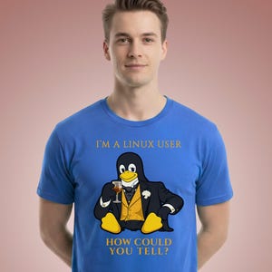 May include: A bright blue short-sleeved t-shirt featuring a graphic of a penguin dressed in a black tuxedo with a yellow vest, holding a glass. The text above reads I'M A LINUX USER and the text below reads HOW COULD YOU TELL? in gold lettering.