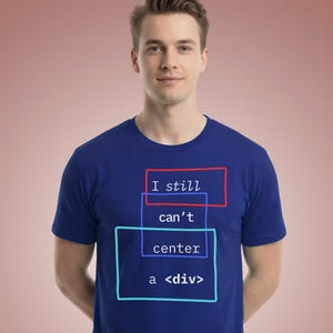 May include: A royal blue short-sleeved t-shirt featuring a graphic design with three overlapping rectangular outlines in red and light blue. The text "I still can't center a <div>" is printed in white, sans-serif font within the rectangular frames.