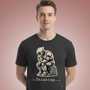 Op de afbeelding: Een zwart t-shirt met korte mouwen en een grafische print van een lichtgekleurde robot die op een rots zit in een houding die doet denken aan het beeld De Denker. Onder de robot staat de tekst Thinking... met een laadicoontje.