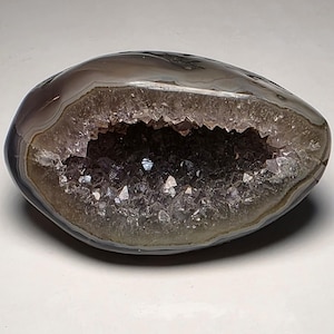 May include: A polished, oval-shaped geode with a dark gray and white banded exterior. The interior cavity is filled with sparkling, light purple amethyst crystals. The geode is displayed against a white background.