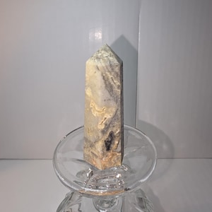 May include: A tall, rectangular crystal obelisk with a pointed top. The stone features a mix of beige, gray, and brown tones. It is displayed on a clear, decorative glass. The obelisk is a natural stone, likely used for decorative or spiritual purposes.