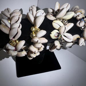 May include: Several seashell bracelets. Each bracelet features multiple white seashells with gold-colored accents. The seashells are arranged in a cluster, creating a unique and natural aesthetic. The bracelets are displayed on a black surface.