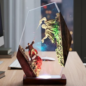 Può includere: Lampada decorativa in resina con una scena di due figure su un paesaggio roccioso. La lampada ha una base in legno e una forma geometrica trasparente. Le figure sono in tonalità di rosso, oro e verde, con un effetto di luce blu.