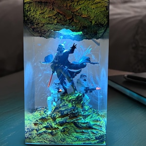 May include: A rectangular resin night light featuring a Star Wars scene with Darth Vader and Stormtroopers. The light has a green and brown textured top, with blue and green lighting illuminating the figures. The lamp is on a dark table.