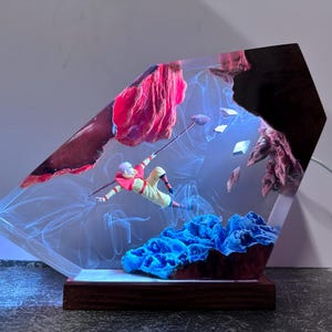May include: A unique resin art piece featuring a figure in a yellow and red outfit, seemingly mid-air, with a staff. The artwork includes blue coral-like structures, red rock formations, and a wooden base. The piece is illuminated with blue light.