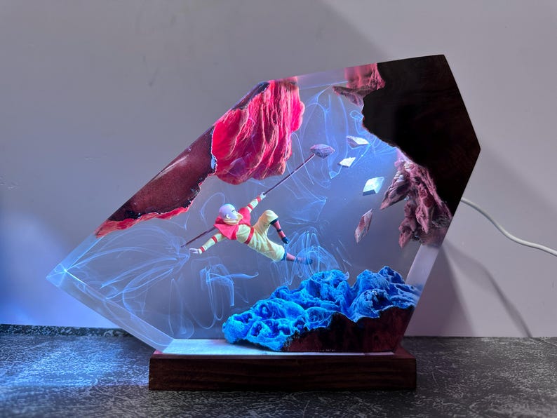 May include: A unique resin art piece featuring a figure in a yellow and red outfit, seemingly mid-air, with a staff. The artwork includes blue coral-like structures, red rock formations, and a wooden base. The piece is illuminated with blue light.