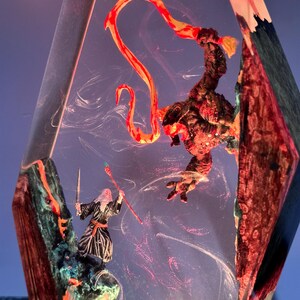 Puede incluir: Escultura de resina de tem&aacute;tica fant&aacute;stica que representa a un mago con espada y b&aacute;culo enfrent&aacute;ndose a un monstruo. La escultura est&aacute; encerrada en un bloque facetado transparente con detalles rojos, naranjas y azules.