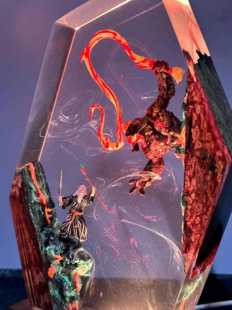 Puede incluir: Escultura de resina de tem&aacute;tica fant&aacute;stica que representa a un mago con espada y b&aacute;culo enfrent&aacute;ndose a un monstruo. La escultura est&aacute; encerrada en un bloque facetado transparente con detalles rojos, naranjas y azules.