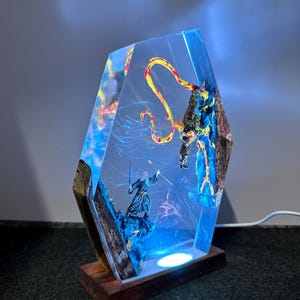 Puede incluir: L&aacute;mpara decorativa de resina con una escena de fantas&iacute;a que representa a un mago y un drag&oacute;n. La l&aacute;mpara tiene una base de madera y un cuerpo de resina transparente y geom&eacute;trico con detalles internos. El interior est&aacute; iluminado con luz azul y blanca.