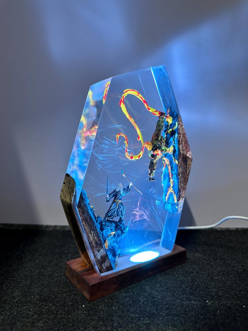 Puede incluir: L&aacute;mpara decorativa de resina con una escena de fantas&iacute;a que representa a un mago y un drag&oacute;n. La l&aacute;mpara tiene una base de madera y un cuerpo de resina transparente y geom&eacute;trico con detalles internos. El interior est&aacute; iluminado con luz azul y blanca.