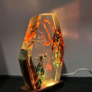 Puede incluir: L&aacute;mpara decorativa que representa a un guerrero luchando contra una criatura, encapsulada en resina transparente con detalles en naranja y verde. La l&aacute;mpara se apoya en una base de madera y tiene una luz c&aacute;lida.