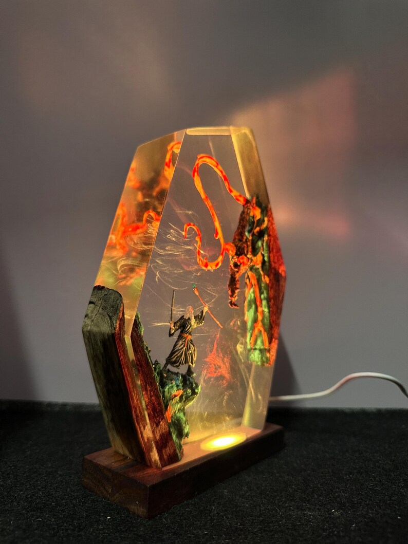 Puede incluir: L&aacute;mpara decorativa que representa a un guerrero luchando contra una criatura, encapsulada en resina transparente con detalles en naranja y verde. La l&aacute;mpara se apoya en una base de madera y tiene una luz c&aacute;lida.