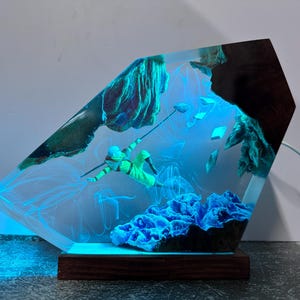 May include: A unique resin art piece featuring a diver in a neon green suit, suspended in a clear, glowing blue environment. The artwork includes blue coral and rock formations, set on a wooden base. The piece is illuminated with a blue light.
