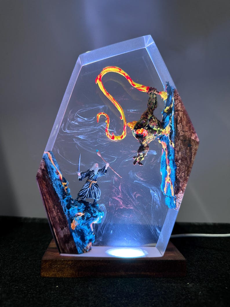 Puede incluir: Una escultura de resina de tem&aacute;tica fant&aacute;stica que representa a un mago luchando contra un drag&oacute;n. La pieza hexagonal transparente tiene una base de madera e iluminaci&oacute;n LED interna. El mago sostiene una espada y un bast&oacute;n, mientras que el drag&oacute;n es rojo y amarillo.