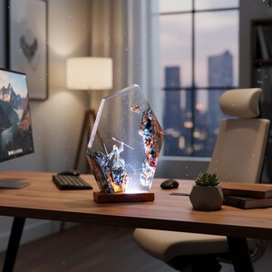 Puede incluir: L&aacute;mpara decorativa de resina con un dise&ntilde;o &uacute;nico. La l&aacute;mpara presenta una escena detallada de un guerrero y un objeto volador dentro de una forma geom&eacute;trica transparente. Se apoya sobre una base de madera y emite un brillo c&aacute;lido, ideal para un escritorio o mesa.