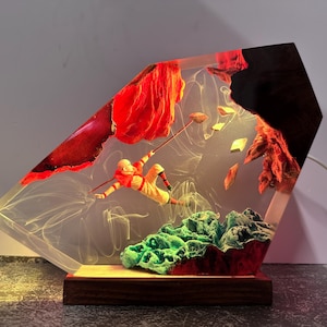 May include: A unique resin art piece featuring a figure in orange and yellow clothing, seemingly climbing with a staff. The artwork includes red and green elements, with a wooden base and a light source, creating a decorative lamp.