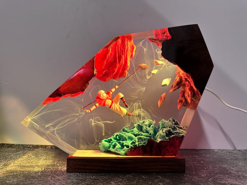 May include: A unique resin art piece featuring a figure in orange and yellow clothing, seemingly climbing with a staff. The artwork includes red and green elements, with a wooden base and a light source, creating a decorative lamp.