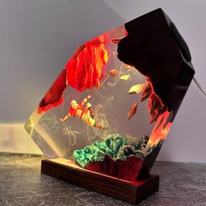 May include: A unique resin lamp with a wood base and accents. The lamp features a figure suspended in the resin, with red and green elements. The lamp has a warm glow and a white power cord.