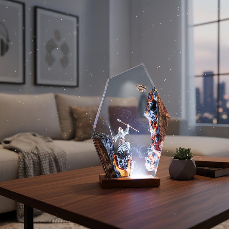 Puede incluir: L&aacute;mpara decorativa que presenta una escena de fantas&iacute;a con una figura de mago, un anillo y un paisaje monta&ntilde;oso encapsulado en resina transparente y geom&eacute;trica. La l&aacute;mpara se asienta sobre una base de madera y emite un brillo c&aacute;lido. La paleta de colores incluye tonos marrones, grises y naranjas.