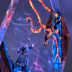 Puede incluir: Una escultura de resina transparente con figuras en miniatura de un mago y una criatura con una llama amarilla y roja. Las figuras se colocan sobre un fondo de patrones arremolinados y coloridos, con tonos azules y rosas.