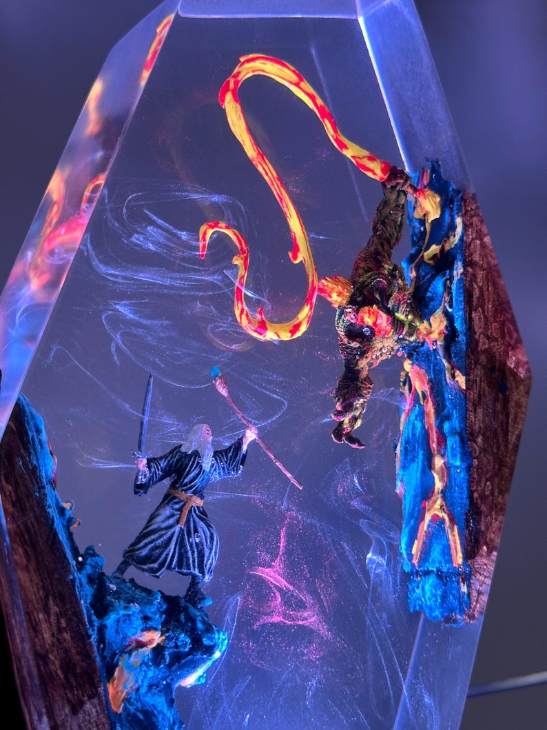 Puede incluir: Una escultura de resina transparente con figuras en miniatura de un mago y una criatura con una llama amarilla y roja. Las figuras se colocan sobre un fondo de patrones arremolinados y coloridos, con tonos azules y rosas.
