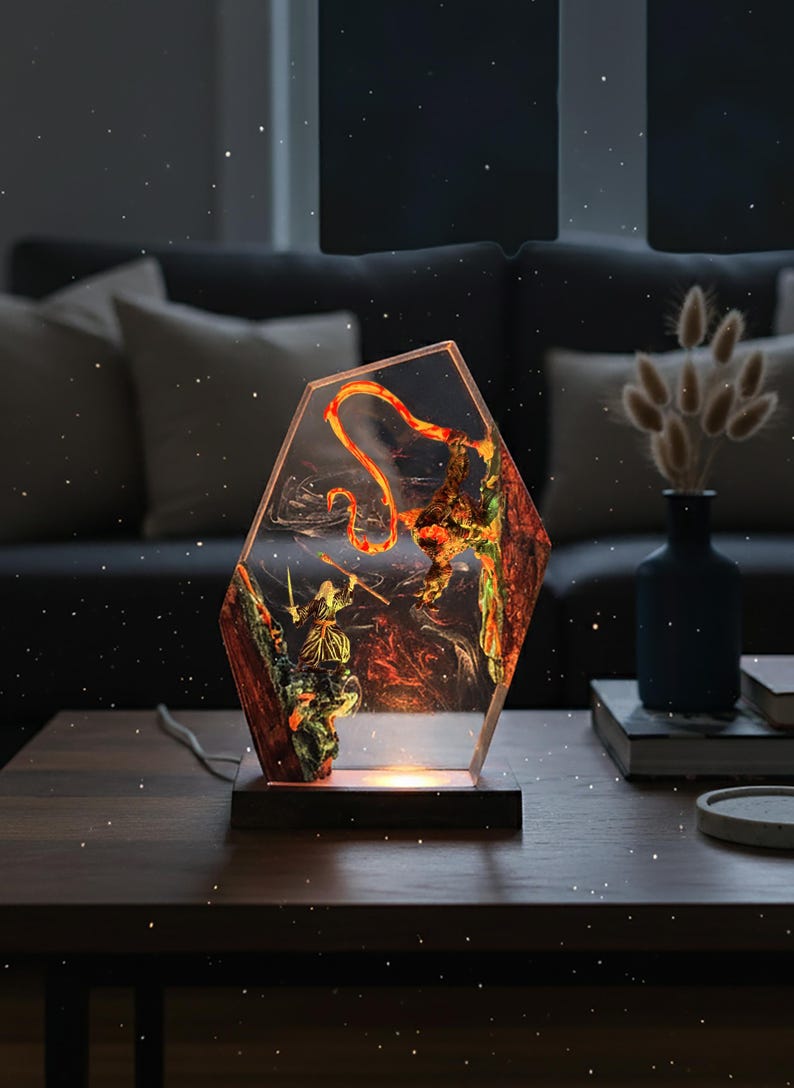 Puede incluir: L&aacute;mpara decorativa con forma hexagonal, con un dise&ntilde;o detallado de resina que representa una figura de guerrero y un drag&oacute;n. La l&aacute;mpara tiene una base de madera y emite un brillo c&aacute;lido y acogedor. La obra de arte interior es una mezcla de rojo, naranja y verde.