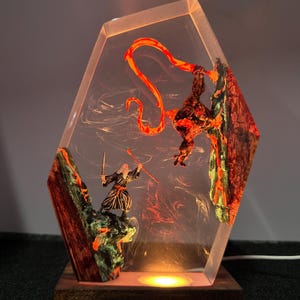 Puede incluir: L&aacute;mpara decorativa con una escena de fantas&iacute;a encapsulada en resina transparente. La escena muestra dos figuras en combate, con un elemento naranja fuego. La l&aacute;mpara tiene una base de madera y emite un brillo c&aacute;lido. La resina tiene forma geom&eacute;trica.